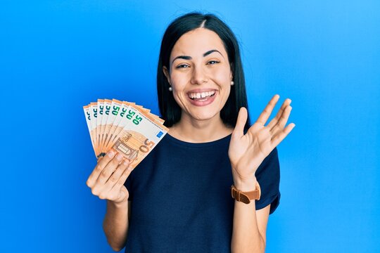 Beautiful young woman holding bunch of 50 euro banknotes waiving saying hello happy and smiling, friendly welcome gesture
