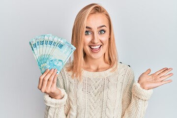 Beautiful caucasian blonde woman holding 100 brazilian real banknotes celebrating achievement with happy smile and winner expression with raised hand