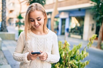 Young blonde girl smiling happy using smartphone at the city.