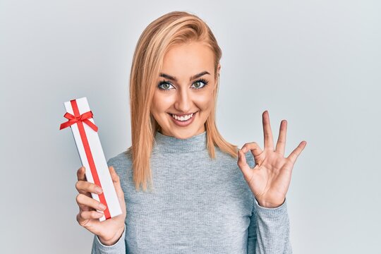 Beautiful caucasian woman holding gift doing ok sign with fingers, smiling friendly gesturing excellent symbol
