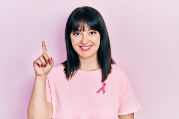 Young hispanic woman wearing pink cancer ribbon on shirt surprised with an idea or question pointing finger with happy face, number one