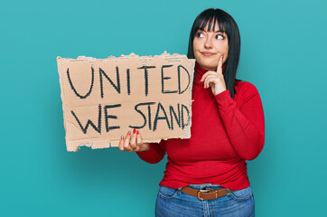 Young hispanic woman holding united we stand banner serious face thinking about question with hand...