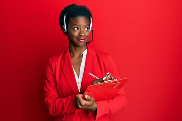 Young african american girl wearing call center agent headset writing on clipboard smiling looking...