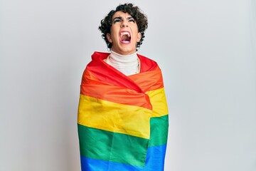 Young man wearing woman make up wrapped in rainbow lgbtq flag angry and mad screaming frustrated and furious, shouting with anger looking up.