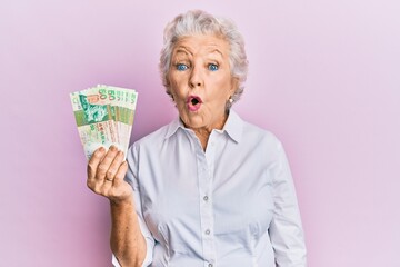 Senior grey-haired woman holding hong kong 50 dollars banknotes scared and amazed with open mouth...