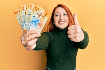 Beautiful redhead woman holding 10 swiss franc banknotes smiling happy and positive, thumb up doing...