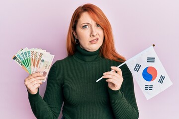 Beautiful redhead woman holding south korean won banknotes and flag in shock face, looking...