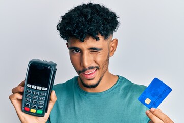 Young arab handsome man holding dataphone and credit card winking looking at the camera with sexy...