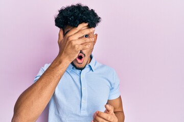 Young arab handsome man wearing casual clothes peeking in shock covering face and eyes with hand,...