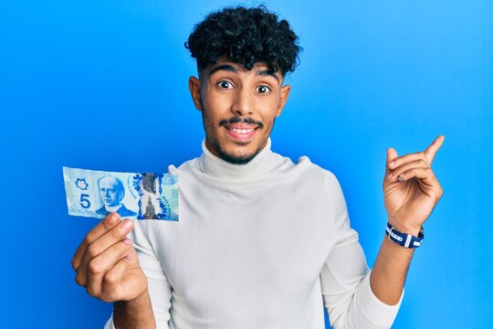 Young arab handsome man holding 5 canadian dollars banknote smiling happy pointing with hand and finger to the side