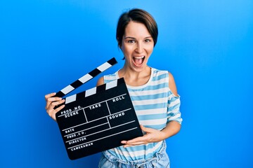 Young brunette woman with short hair holding video film clapboard celebrating crazy and amazed for...