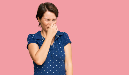 Young brunette woman with short hair wearing casual clothes smelling something stinky and disgusting, intolerable smell, holding breath with fingers on nose. bad smell