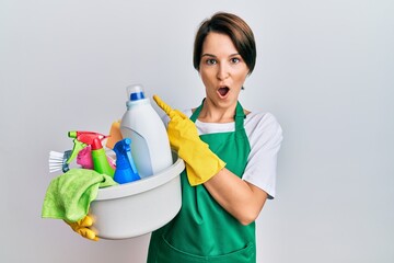 Young brunette woman with short hair wearing apron holding cleaning products surprised pointing with finger to the side, open mouth amazed expression.
