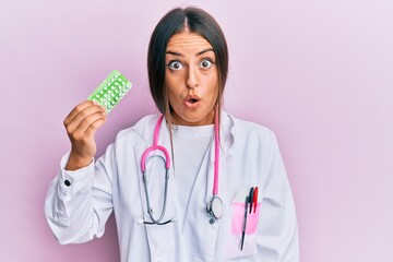 Beautiful hispanic woman wearing doctor uniform holding prescription pills scared and amazed with...