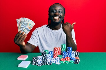 Handsome young black man playing poker holding 100 dollars banknotes pointing thumb up to the side smiling happy with open mouth
