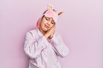 Obraz na płótnie Canvas Hispanic woman with pink hair wearing sleep mask and pajama sleeping tired dreaming and posing with hands together while smiling with closed eyes.