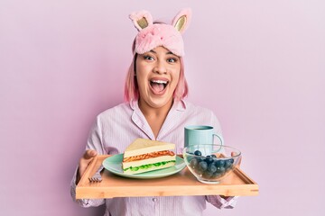 Hispanic woman with pink hair wearing pajama holding healthy breakfast celebrating crazy and amazed for success with open eyes screaming excited.