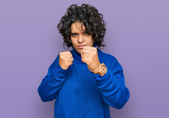 Young hispanic woman with curly hair wearing turtleneck sweater ready to fight with fist defense gesture, angry and upset face, afraid of problem