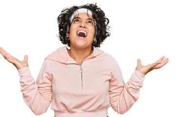 Young hispanic woman with curly hair wearing sportswear crazy and mad shouting and yelling with aggressive expression and arms raised. frustration concept.