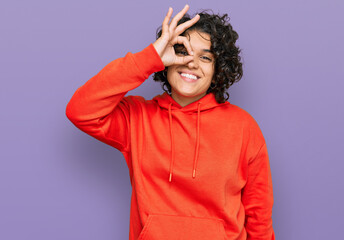 Young hispanic woman with curly hair wearing casual sweatshirt smiling happy doing ok sign with hand on eye looking through fingers