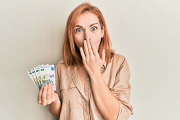 Young caucasian woman holding bunch of 5 euro banknotes covering mouth with hand, shocked and...