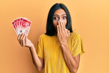 Young brunette woman holding 20 israel shekels banknotes covering mouth with hand, shocked and...