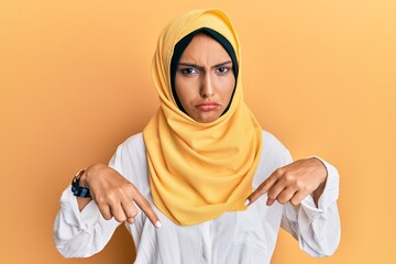 Young brunette arab woman wearing traditional islamic hijab scarf pointing down looking sad and upset, indicating direction with fingers, unhappy and depressed.