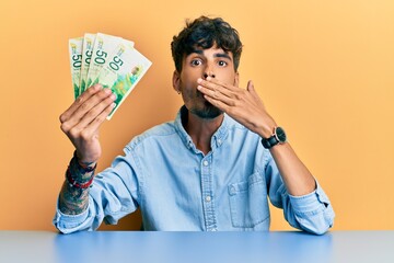 Young hispanic man holding israel shekels sitting on the table covering mouth with hand, shocked and afraid for mistake. surprised expression