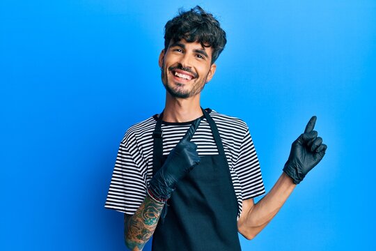 Young hispanic man wearing barber apron smiling and looking at the camera pointing with two hands and fingers to the side.