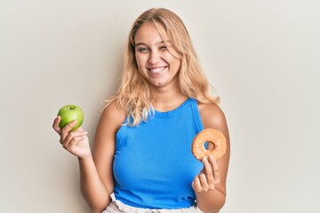 Young blonde girl holding green apple and donut winking looking at the camera with sexy expression,...