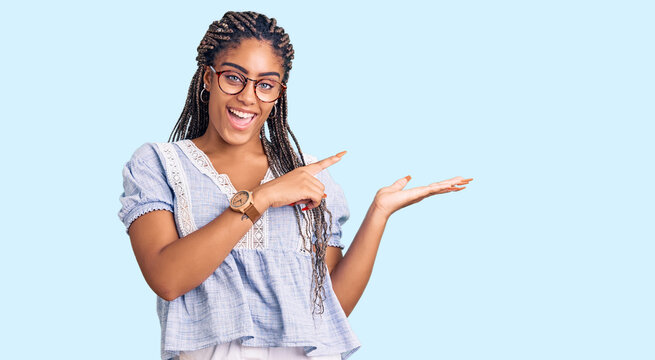 Young african american woman with braids wearing casual summer clothes and glasses amazed and smiling to the camera while presenting with hand and pointing with finger.