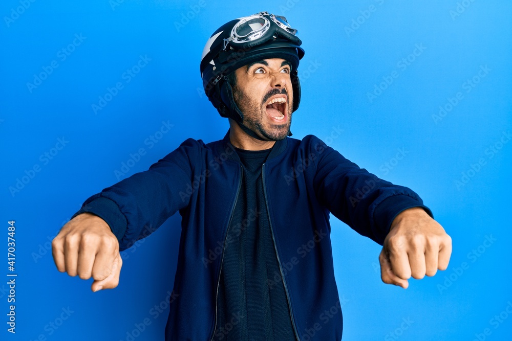 Poster young hispanic man holding motorcycle helmet angry and mad screaming frustrated and furious, shoutin