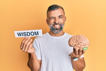 Middle age handsome man holding wisdom word on paper and brain winking looking at the camera with...