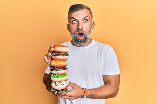 Middle age handsome man holding tasty colorful doughnuts afraid and shocked with surprise and amazed expression, fear and excited face.