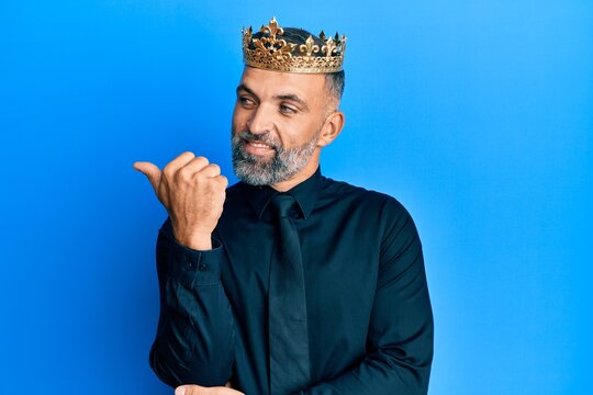 Middle age handsome business man wearing king crown smiling with happy face looking and pointing to the side with thumb up.