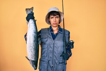 Beautiful brunettte fisher woman holding fishing rod and raw salmon skeptic and nervous, frowning...