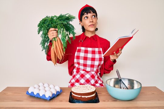 Beautiful brunettte woman cooking carrot cake reading recipes book smiling looking to the side and staring away thinking.