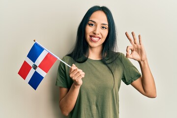 Young hispanic girl holding dominican republic flag doing ok sign with fingers, smiling friendly gesturing excellent symbol - obrazy, fototapety, plakaty