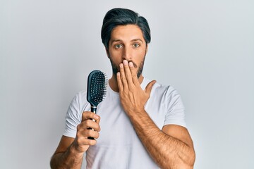 Young hispanic man styling hair using comb covering mouth with hand, shocked and afraid for...