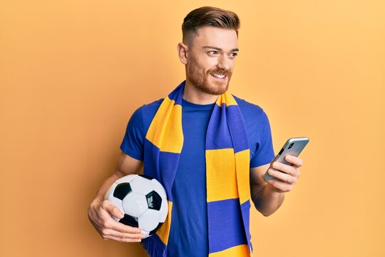 Young redhead man wearing hooligan scarf and using smartphone smiling looking to the side and staring away thinking.