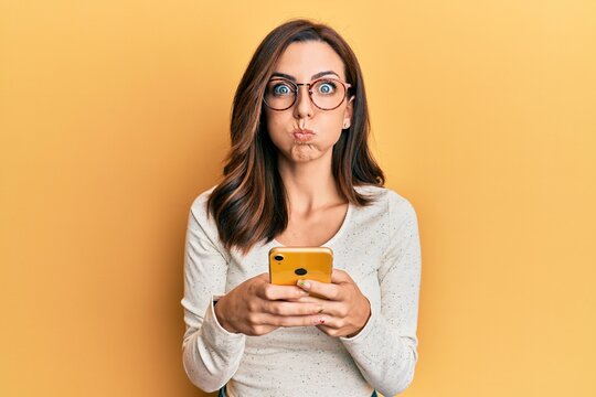 Young brunette woman using smartphone over yellow background puffing cheeks with funny face. mouth inflated with air, catching air.
