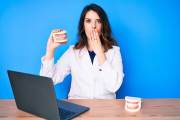 Young beautiful brunette woman working at dentist clinic holding denture covering mouth with hand,...