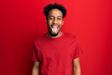 Young african american man with beard wearing casual red t shirt sticking tongue out happy with funny expression. emotion concept.