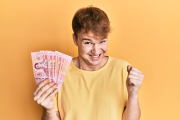 Young caucasian man holding 100 thai baht banknotes screaming proud, celebrating victory and success very excited with raised arm