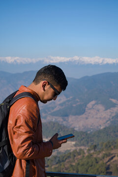 picture of boy using his smartphone on top of mountain. boy wearing brown leather coat and a bagpack