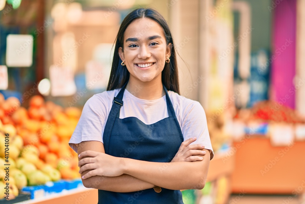 Sticker Young latin shopkeeper girl with arms crossed smiling happy at the fruit store.