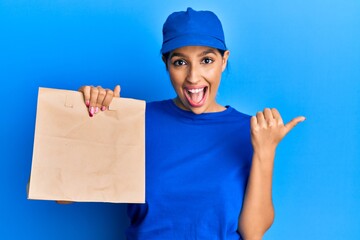 Beautiful brunette woman holding take away paper bag pointing thumb up to the side smiling happy with open mouth