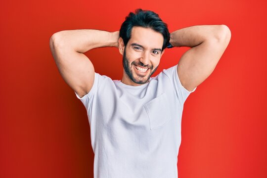 Young hispanic man wearing casual white tshirt relaxing and stretching, arms and hands behind head and neck smiling happy