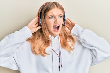 Beautiful young caucasian girl listening to music using headphones angry and mad screaming frustrated and furious, shouting with anger. rage and aggressive concept.