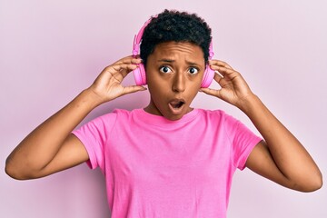 Young african american girl listening to music using headphones in shock face, looking skeptical and sarcastic, surprised with open mouth